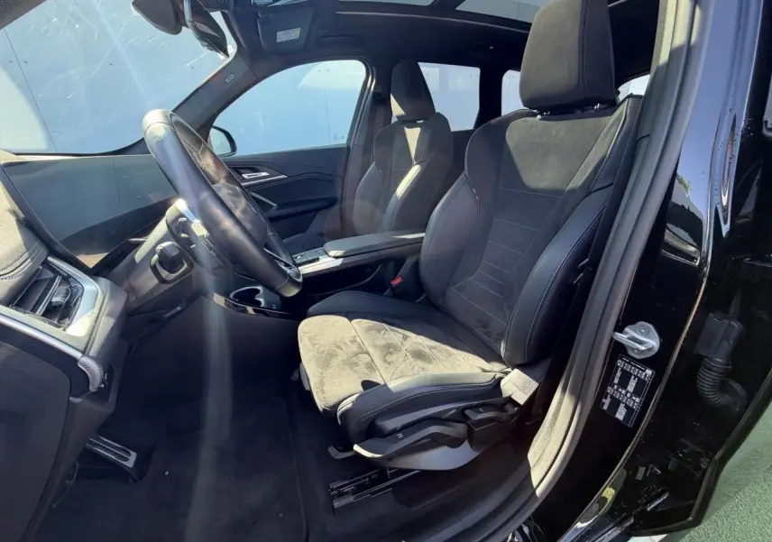 Vue intérieure côté conducteur du BMW X1 noir saphir, sièges sport en Alcantara avec surpiqûres bleues et toit panoramique visible.