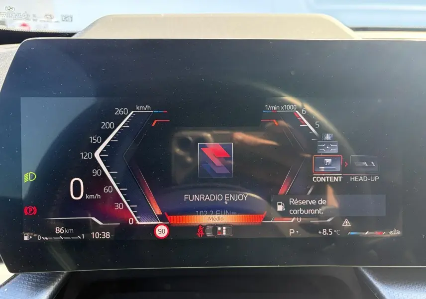 Gros plan sur le combiné d'instrumentation numérique du BMW X1 noir, affichant la vitesse, la radio et l'alerte réserve de carburant.