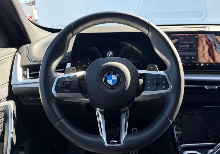 Vue frontale du volant cuir M noir avec commandes intégrées, tableau de bord numérique et écran tactile du BMW X1 noir Saphir 2025.