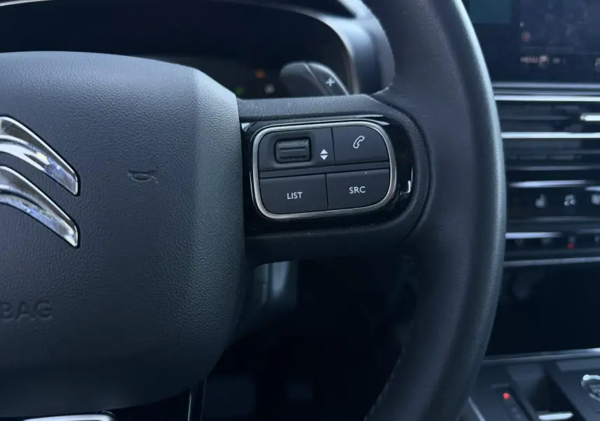 Gros plan sur les commandes gauche du volant cuir noir du Citroën C5 Aircross Hybrid 2025, avec tableau de bord en arrière-plan.