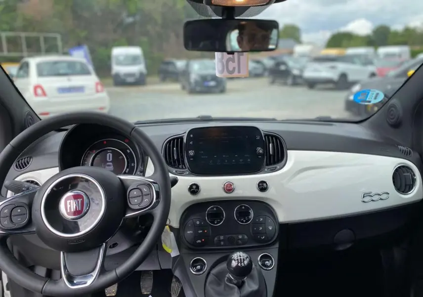 Vue intérieure avant du tableau de bord blanc et noir d'une Fiat 500 2024 avec volant multifonction et boîte manuelle.