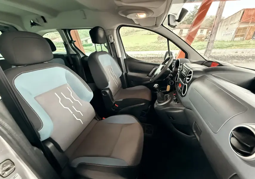 Vue intérieure côté conducteur du Peugeot Partner Tepee 2014, sièges tissu gris et bleu avec volant cuir noir.