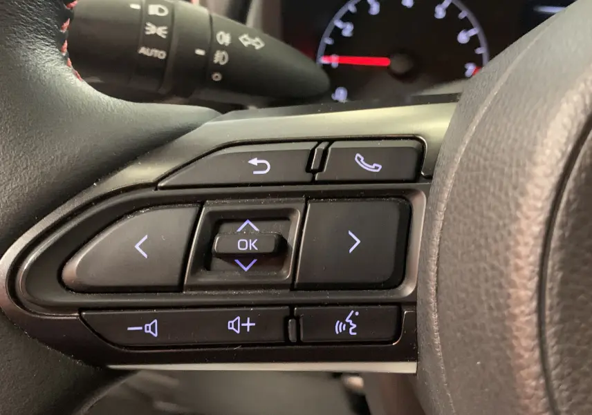 Gros plan sur les commandes au volant côté gauche de la Toyota Yaris blanche, avec boutons de navigation et téléphone.