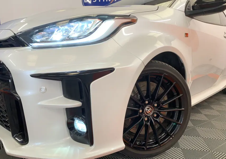 Gros plan sur l'avant droit blanc métal de la Toyota Yaris GR 1.6 VVT-I avec phares LED allumés et jantes noires sport.