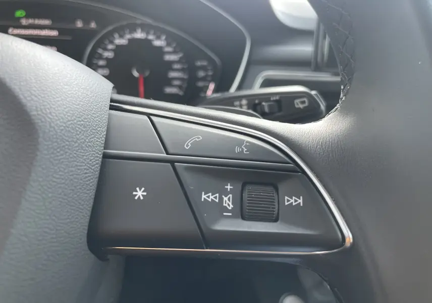 Gros plan sur les commandes audio au volant d'une Audi A4 Avant 40 TDI blanc, avec tableau de bord flou en arrière-plan.