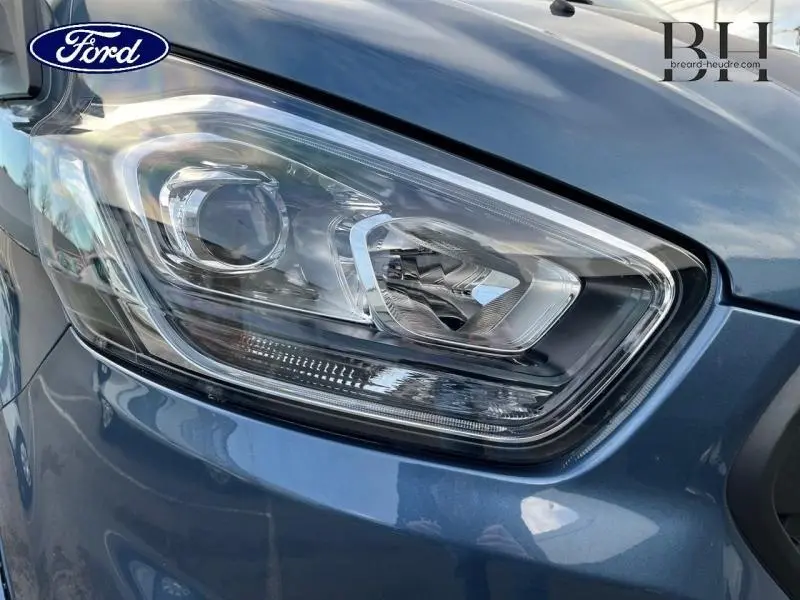 Gros plan sur le phare avant gauche du Ford Transit Custom bleu azur métallisé, mettant en valeur son design moderne.