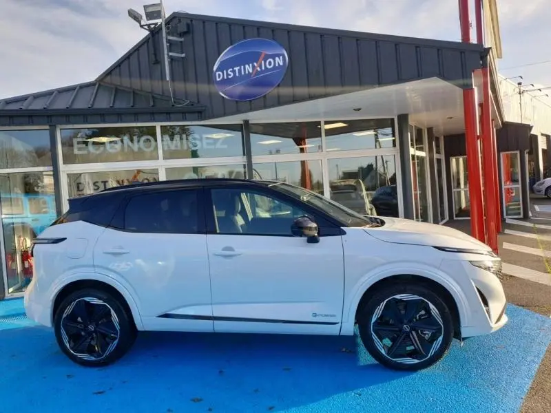 Vue de profil côté gauche d'un Nissan Qashqai Hybrid e-POWER 2025 blanc nacré avec toit noir devant un showroom.