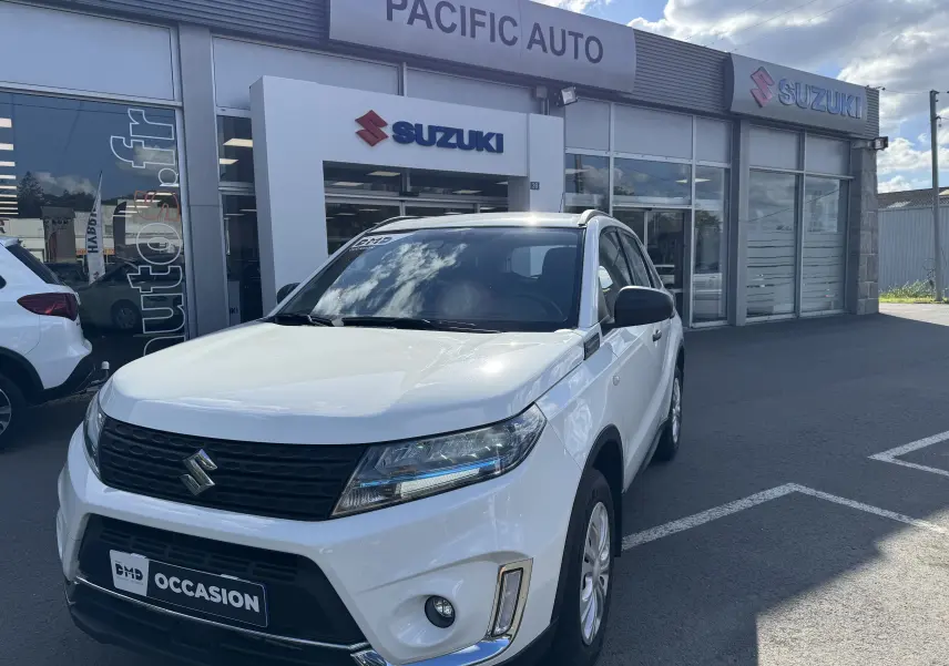 Vue 3/4 avant d'un Suzuki Vitara blanc hybride 2023 garé devant un concessionnaire Suzuki avec logo visible.