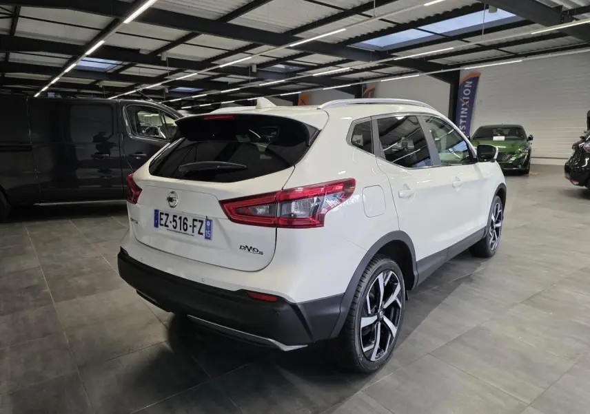 Vue 3/4 arrière droite d'un Nissan Qashqai blanc avec jantes alliage noires et toit panoramique en intérieur showroom.