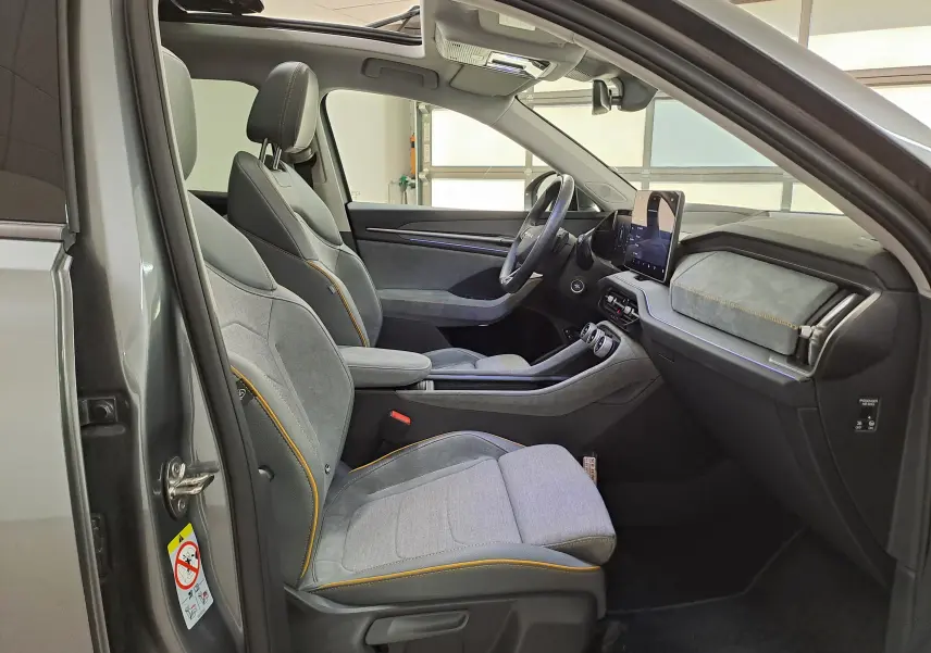 Vue intérieure côté conducteur du Skoda Kodiaq gris graphite 2025, sièges tissu gris avec surpiqûres orange et toit panoramique ouvert.