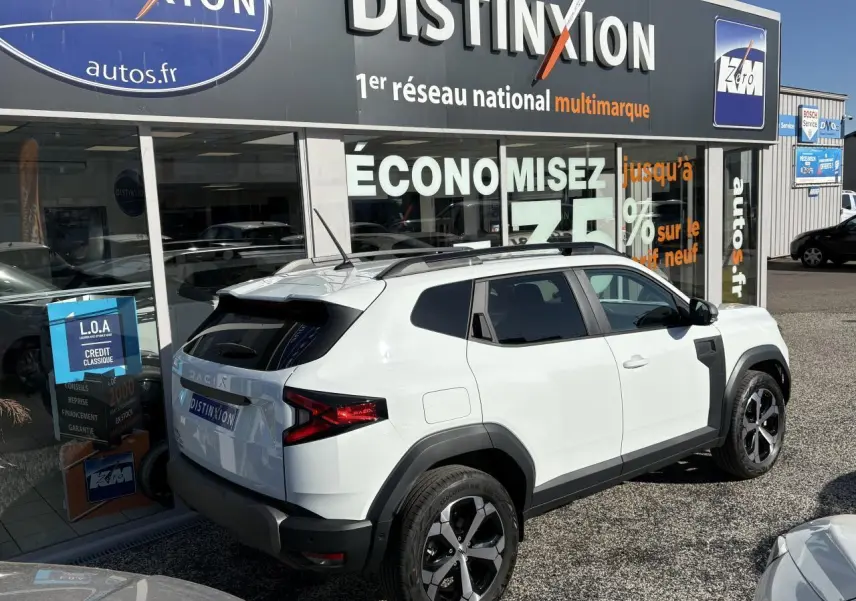 Vue 3/4 arrière droite d'un Dacia Duster 2024 blanc métallisé avec barres de toit noires et jantes alliage bicolores.