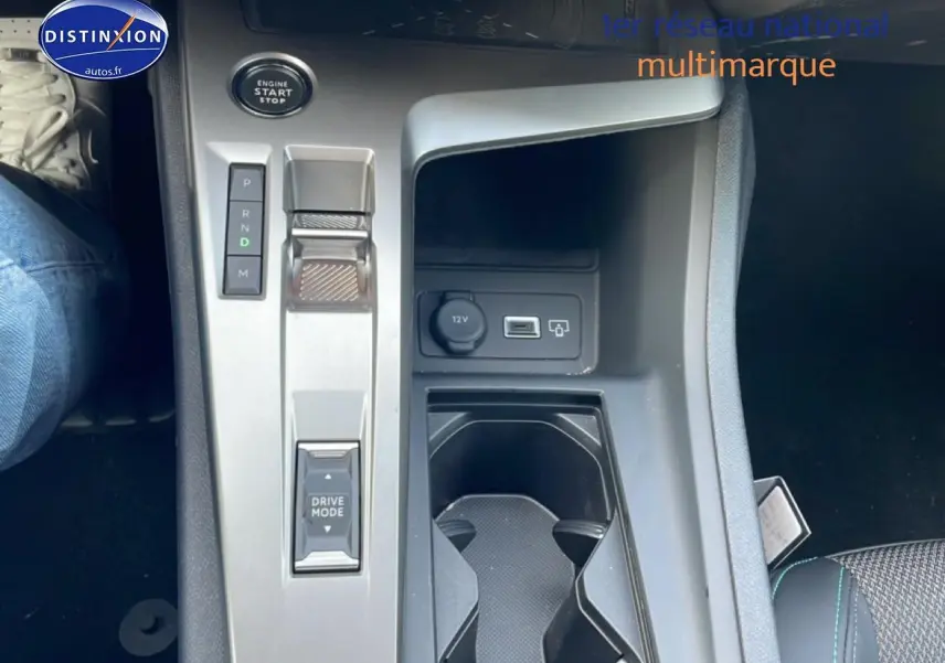 Vue plongeante sur la console centrale grise de la Peugeot 308 hybride 2025 avec bouton start et prise 12V visible.