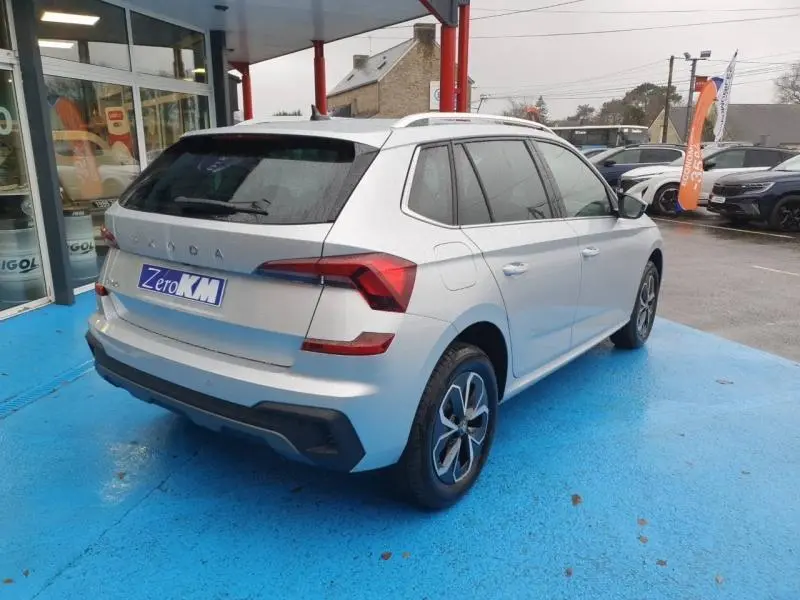 Vue 3/4 arrière droite d'un Skoda Kamiq gris diamant métallisé avec barres de toit et jantes alu au showroom.