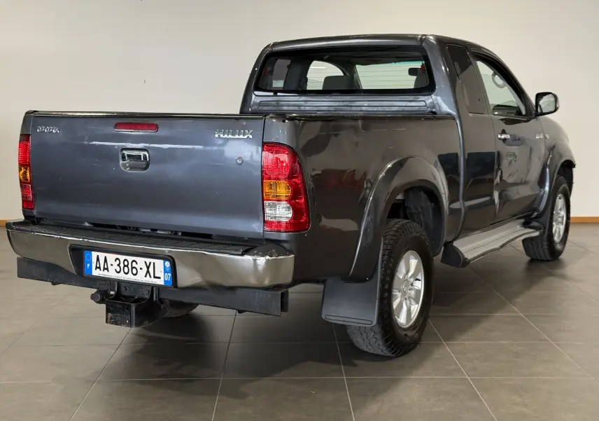 Vue 3/4 arrière droite d'un Toyota Hilux X-Tra Cabine gris 2009 avec benne et pare-chocs arrière légèrement abîmé.