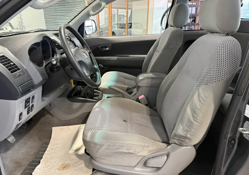 Vue intérieure côté conducteur du Toyota Hilux X-Tra Cabine 2009, montrant sièges gris et tableau de bord avec boîte manuelle.