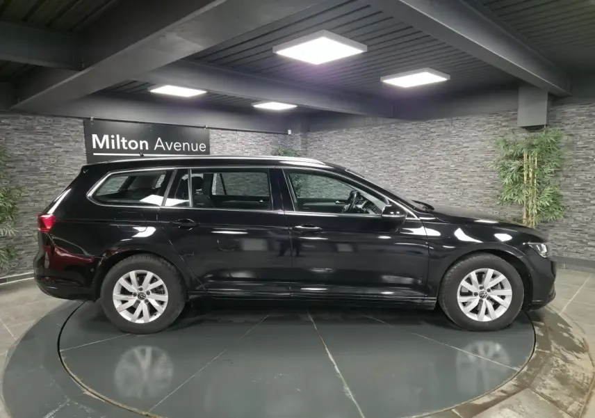 Volkswagen Passat SW noir vue de profil côté gauche dans un showroom avec sol gris et mur en pierre.