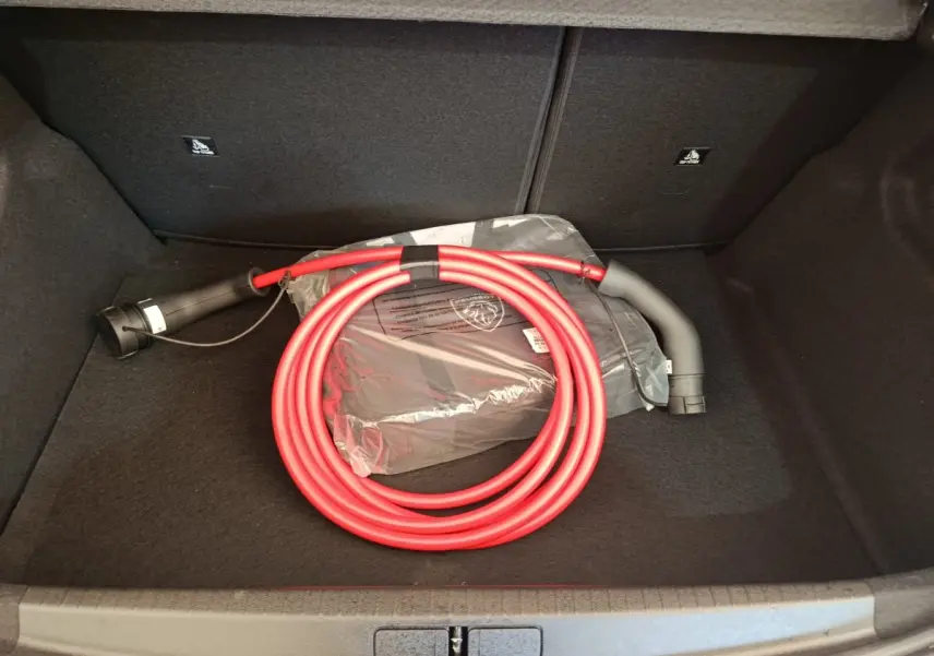 Coffre ouvert d’une Peugeot 208 électrique 2024 avec câble de charge rouge enroulé et sac de rangement noir.