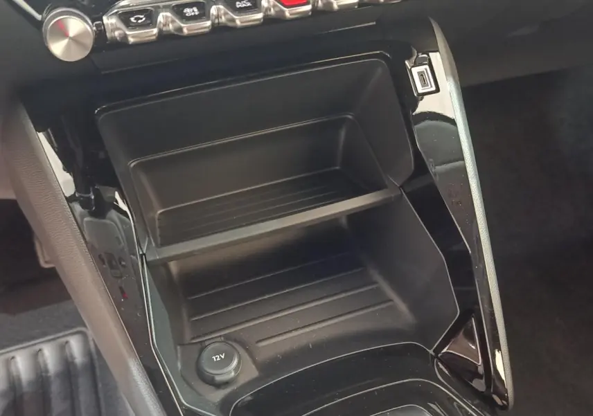 Gros plan sur la console centrale noire de la Peugeot 208 électrique 2024 avec prise 12V et rangement intégré.