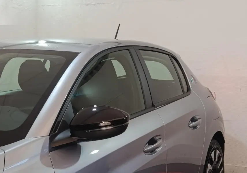 Vue côté gauche rapprochée d’une Peugeot 208 électrique gris clair avec rétroviseur noir brillant et vitres teintées.