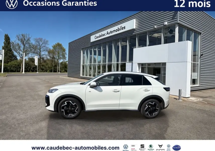 Volkswagen T-Roc blanc vu de profil côté gauche, avec jantes noires et bâtiment de concession en arrière-plan.