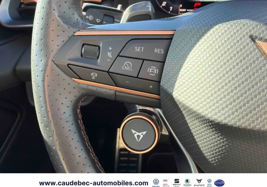 Gros plan sur le volant perforé noir avec commandes et logo CUPRA cuivré, tableau de bord partiellement visible.