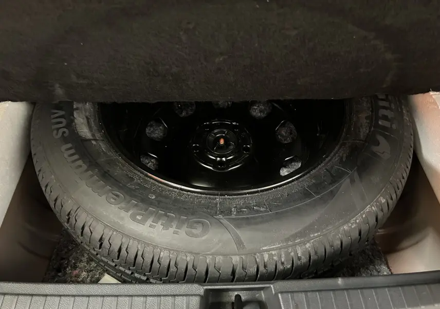 Roue de secours noire dans le coffre du Dacia Duster orange Arizona, vue rapprochée sous la moquette du coffre.