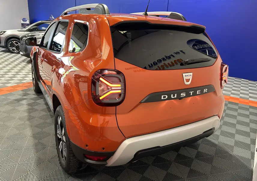 Vue 3/4 arrière droite du Dacia Duster orange Arizona 2022 avec feux arrière allumés et barres de toit visibles.