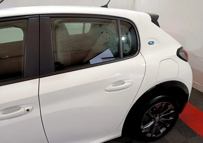 Vue latérale côté droit de la Peugeot 208 électrique blanche, mettant en valeur la porte arrière et le logo "e" bleu près de l'aile arrière.