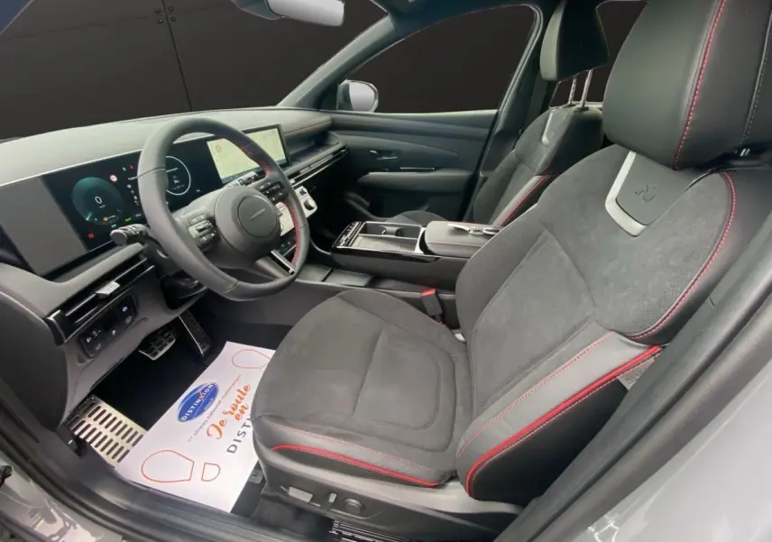 Vue intérieure côté conducteur du Hyundai Tucson 1.6 Hybrid gris foncé, sièges noirs avec surpiqûres rouges et tableau de bord numérique.