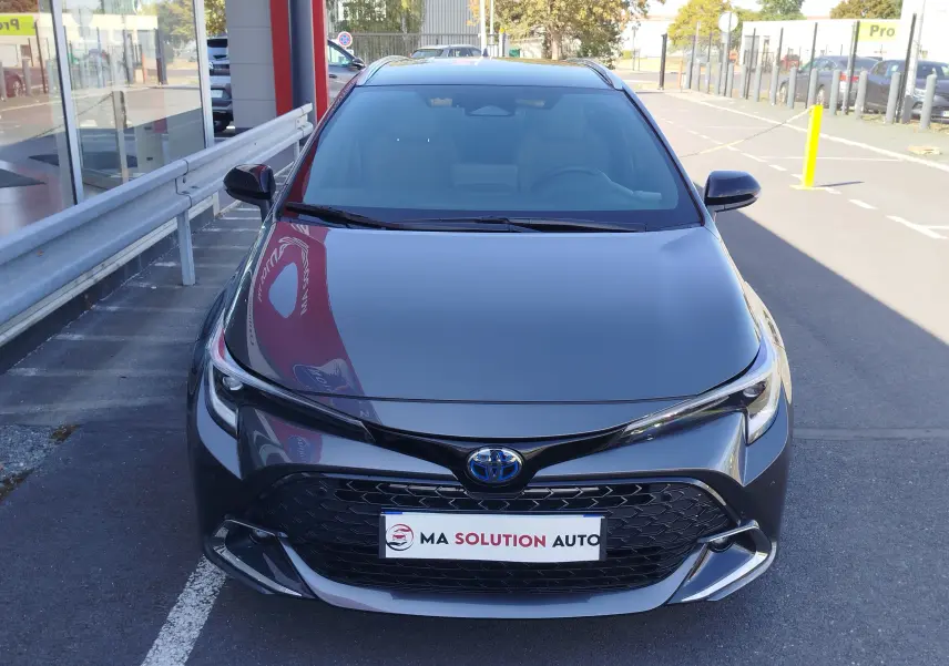 Vue avant frontale d'une Toyota Corolla Touring Sport gris atlas avec calandre noire et logo bleu hybride visible.