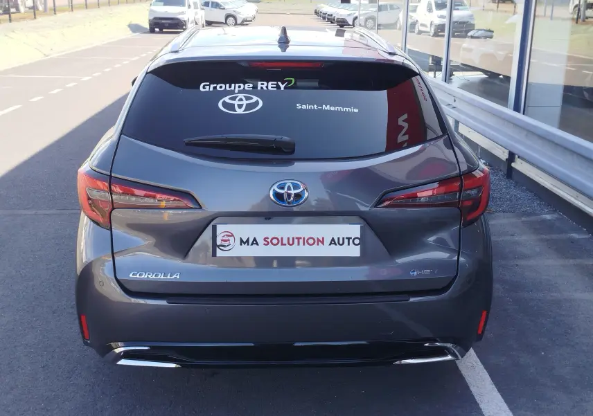 Vue arrière d'une Toyota Corolla Touring Sport gris atlas avec logo hybride et vitres teintées, stationnée devant un bâtiment.