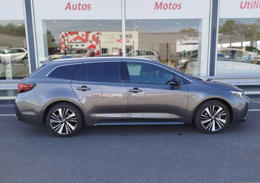 Profil latéral droit d'une Toyota Corolla Touring Sport gris Atlas avec jantes alliage bicolores, stationnée devant une vitrine.