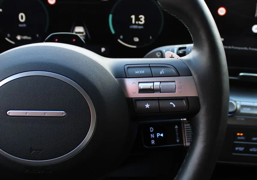 Gros plan sur le volant noir cuir du Hyundai Kona 2024, avec commandes intégrées et tableau de bord numérique en arrière-plan.