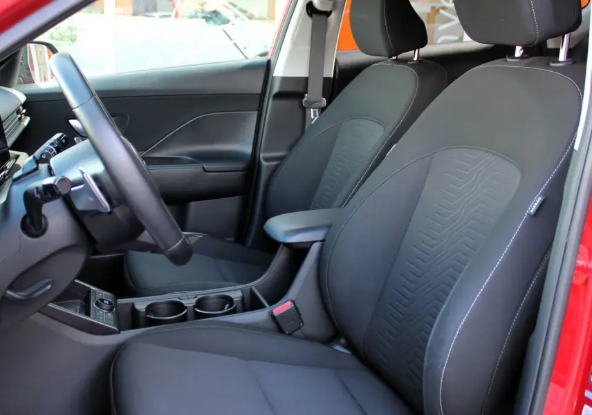 Vue intérieure côté conducteur du Hyundai Kona 2024 rouge, sièges avant noirs en tissu avec surpiqûres blanches.