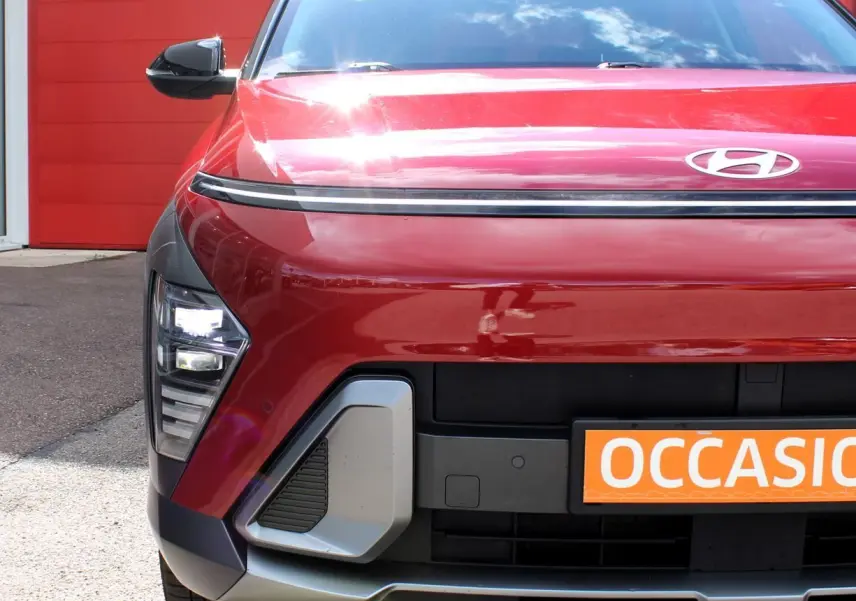 Vue avant rapprochée d’un Hyundai Kona rouge Ultimate Metal avec calandre noire et détails gris, devant un mur rouge.