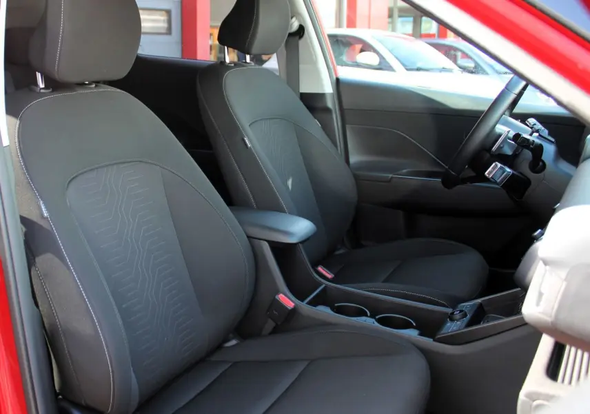 Vue intérieure côté gauche du Hyundai Kona 2024 hybride rouge, montrant les sièges avant en tissu noir avec accoudoir central.
