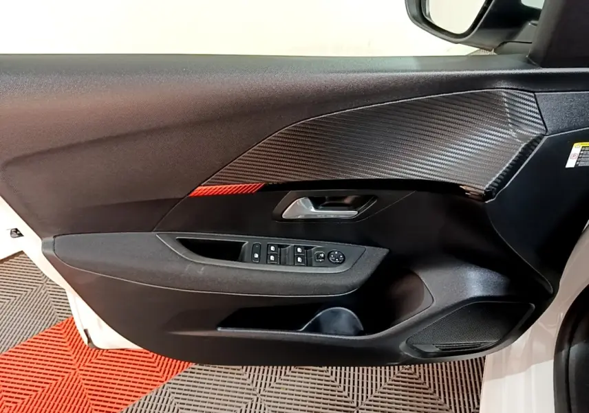 Vue intérieure du panneau de porte avant gauche de la Peugeot 208 électrique blanche, avec commandes de vitres et insert effet carbone.