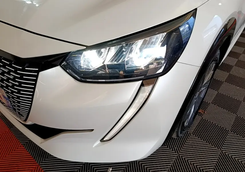 Gros plan sur l’avant droit blanc d’une Peugeot 208 électrique 2021, mettant en valeur le phare LED et la calandre distinctive.