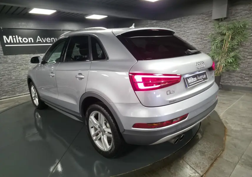 Audi Q3 gris métallisé vue 3/4 arrière droit avec feux arrière allumés dans un showroom intérieur.