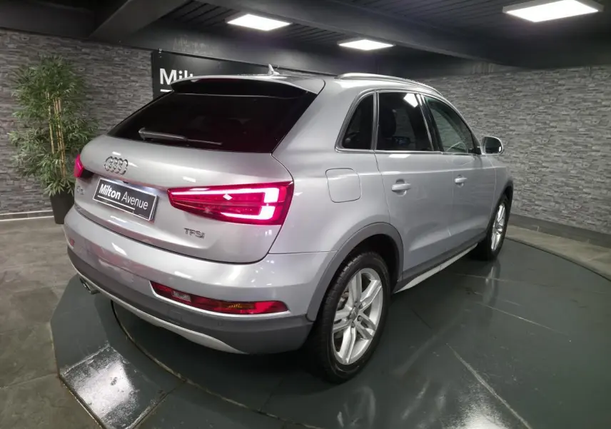 Audi Q3 gris métallisé vue 3/4 arrière droit avec feux arrière LED allumés en intérieur showroom.