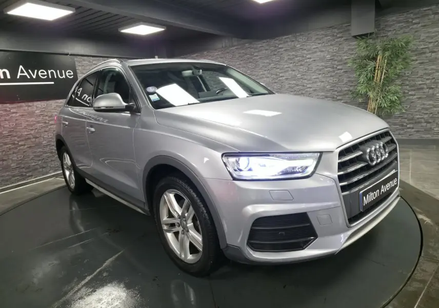 Audi Q3 gris métallisé en 3/4 avant droit, avec calandre Singleframe et phares LED allumés dans un showroom.
