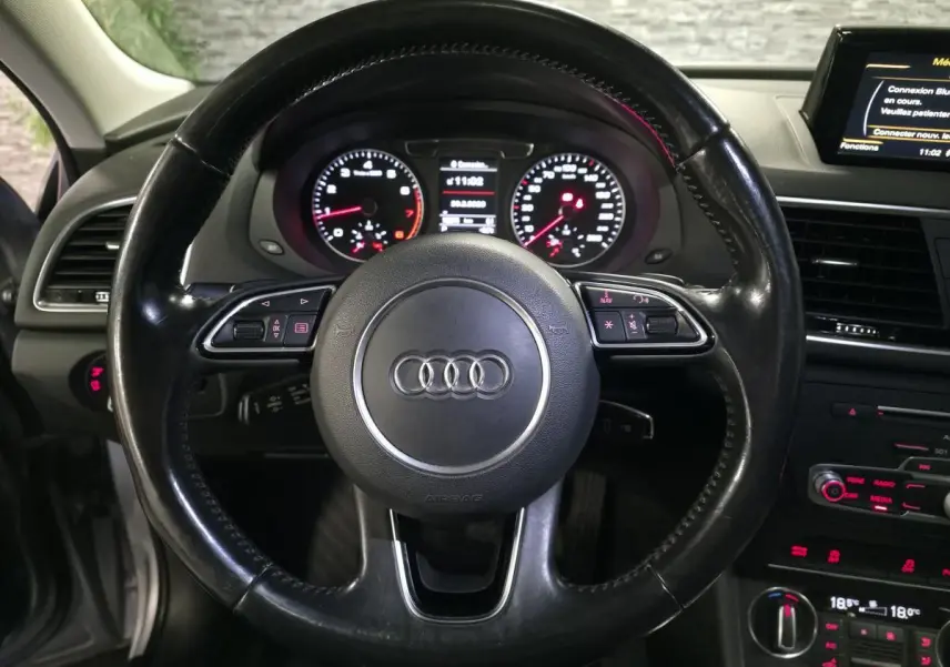 Vue intérieure centrée sur le volant en cuir noir avec logo Audi, tableau de bord et console centrale du Q3 gris de 2016.