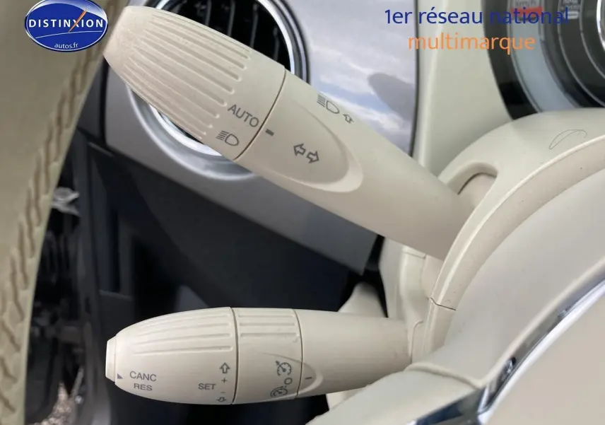Gros plan sur les commandes blanches du volant d’une FIAT 500 gris, mettant en valeur les fonctions de régulateur et feux automatiques.