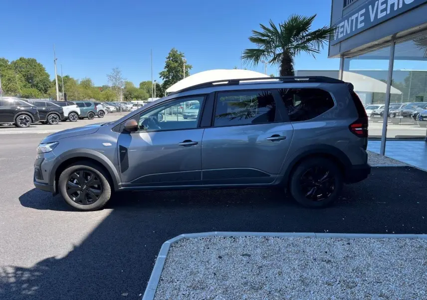 Profil droit du Dacia Jogger 1.6 Hybrid gris schiste 2025 avec jantes noires et toit noir, stationné devant un showroom.