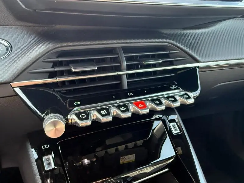 Vue rapprochée de la console centrale noire brillante du Peugeot 2008 Hybrid 2025 avec boutons métalliques et finition carbone.