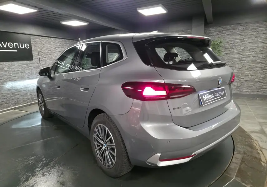 BMW Série 2 218d Active Tourer gris métallisé vue 3/4 arrière avec feux arrière LED allumés en intérieur showroom.