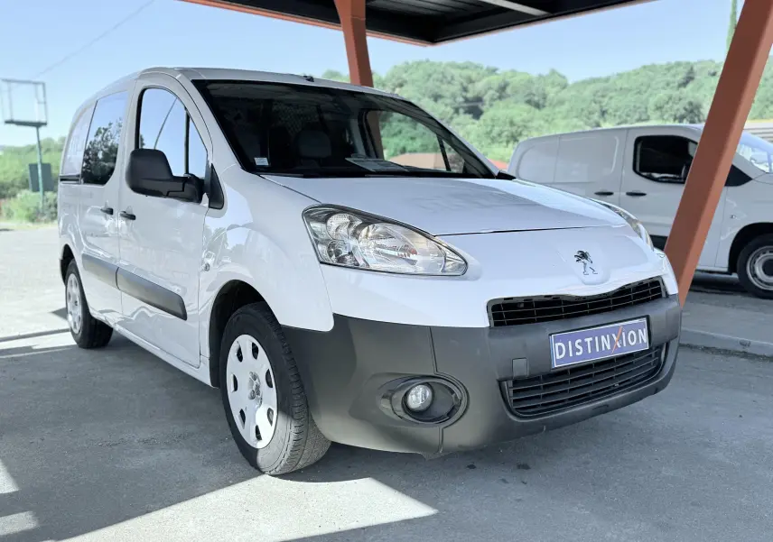 Peugeot Partner blanc vu en 3/4 avant droit, avec pare-chocs noirs et jantes acier sous un abri.