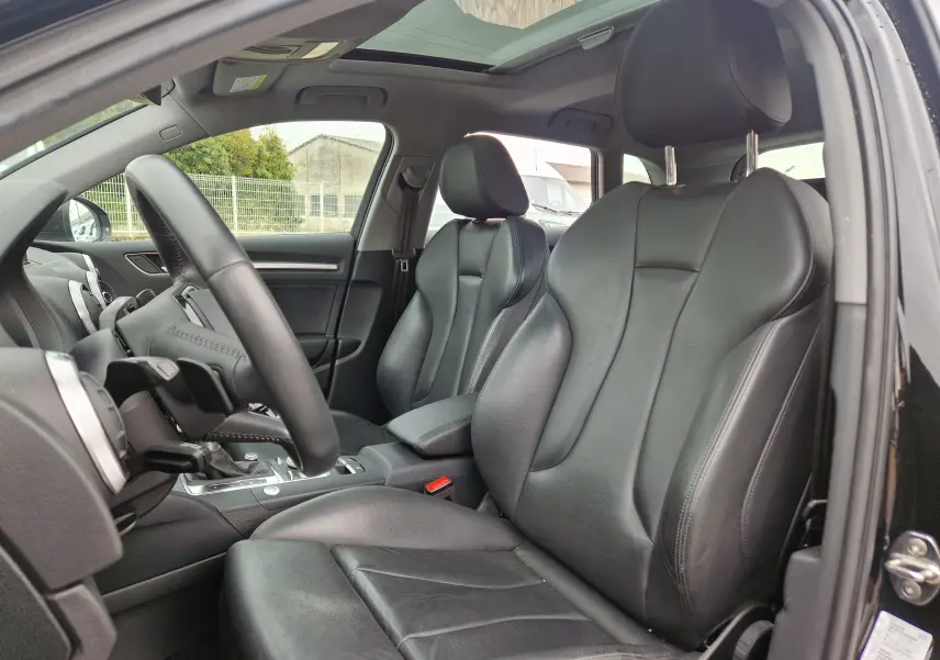 Vue intérieure côté conducteur de l'Audi A3 Sportback noir, sièges en cuir noir et toit ouvrant panoramique visibles.