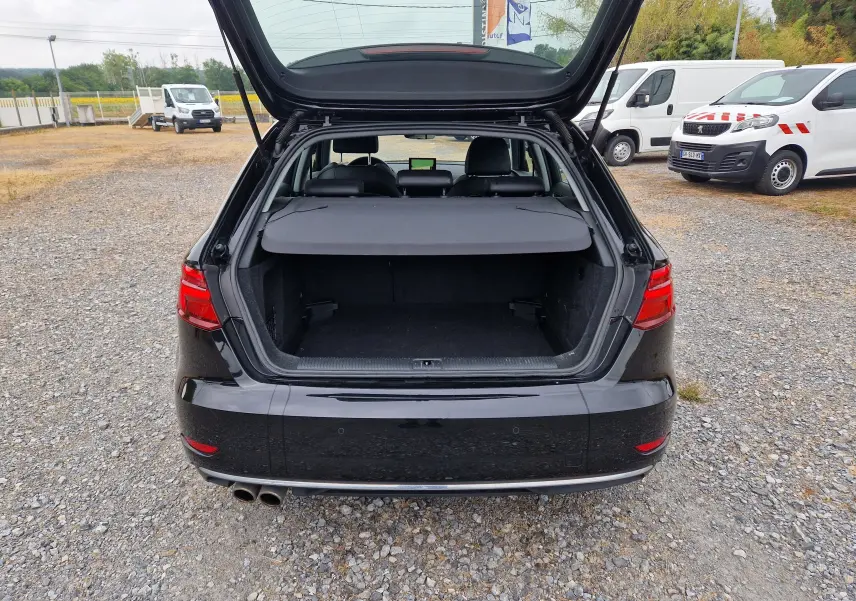 Vue arrière d'Audi A3 Sportback noire 2019 avec coffre ouvert et cache-bagages en place sur un sol gravillonné.