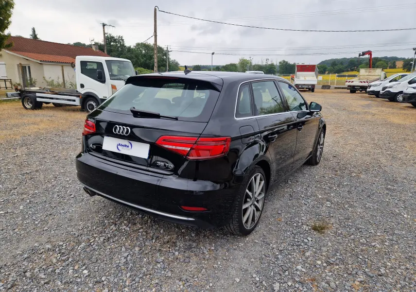 Audi A3 Sportback noir vue 3/4 arrière droit sur un sol gravillonné avec plusieurs véhicules en arrière-plan.